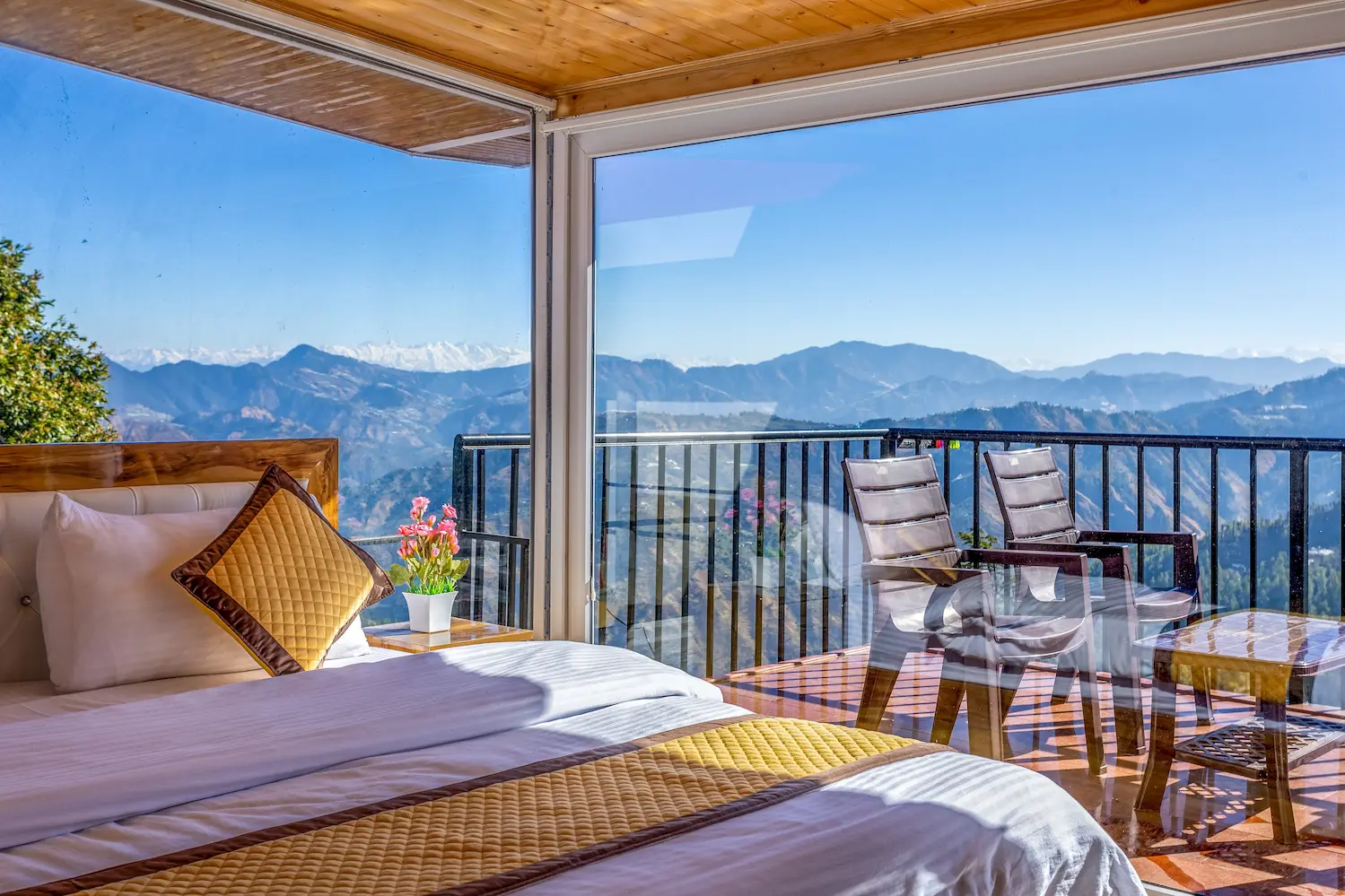 Bed beside floor-to-ceiling glass, with balcony chairs and the far snow line in view